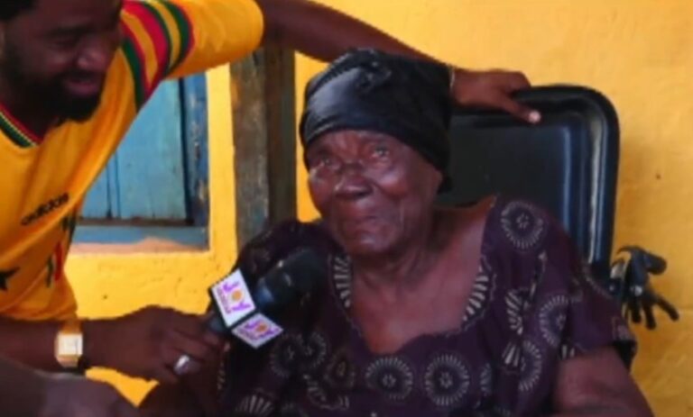 A man interviews an elderly woman in a wheelchair, holding a microphone to her as she sits indoors against a yellow wall.