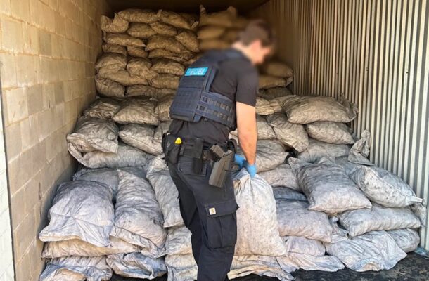 Police officer in tactical vest and gloves stands amid many gray sacks stacked in a metal storage container, inspecting the bags.