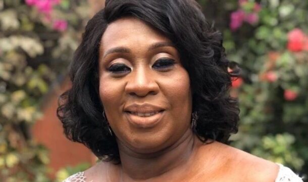 Smiling Black woman with short dark hair and makeup, outdoors with colorful flowers in the background.
