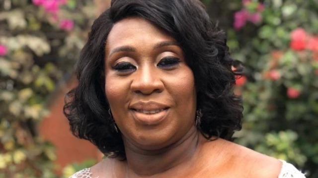 Smiling Black woman with short dark hair and makeup, outdoors with colorful flowers in the background.
