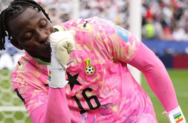 Ghana goalkeeper in pink jersey number 16, touching his gloved fist to his lips in celebration near the goal with a crowd in the background.