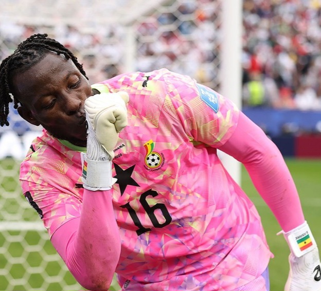 Ghana goalkeeper in pink jersey number 16, touching his gloved fist to his lips in celebration near the goal with a crowd in the background.