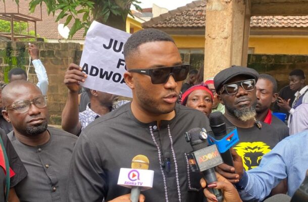 A man in black traditional attire and sunglasses speaks to reporters holding microphones at a protest or press event, with a sign in the background.