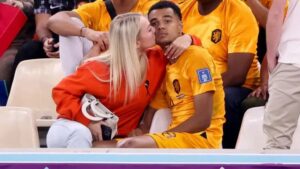 Blonde woman in an orange top kisses a man in a yellow sports jersey on stadium seating.