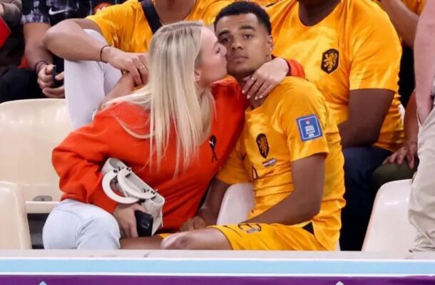 Blonde woman in an orange top kisses a man in a yellow sports jersey on stadium seating.