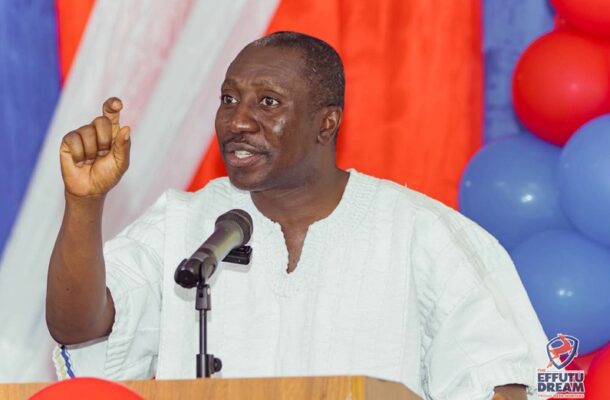 Man delivering a speech at a podium with a microphone, gesturing with his right hand, colorful backdrop with red, white, and blue balloons behind him.