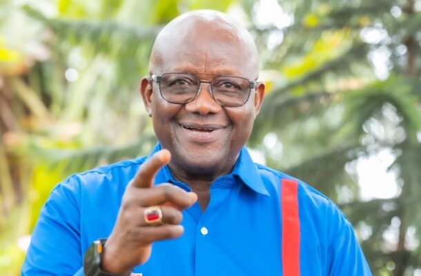Smiling man wearing glasses in a bright blue shirt points toward the camera outdoors.
