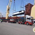 Crane hoists a white passenger train car onto a cargo barge beside a large ship, with a red truck and workers nearby at the port.