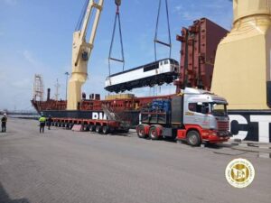 Crane hoists a white passenger train car onto a cargo barge beside a large ship, with a red truck and workers nearby at the port.