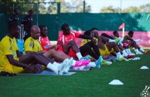 Soccer players in yellow and red jerseys sit on mats doing core stretches during a training session on a green field with a corner flag in the background.