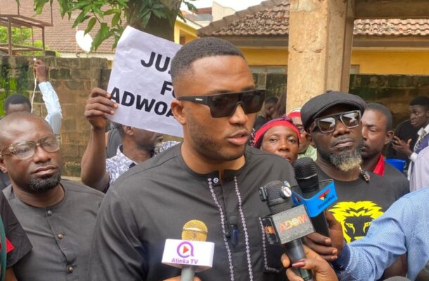 Man wearing dark outfit and sunglasses speaks to reporters at a protest, microphones in front of him.