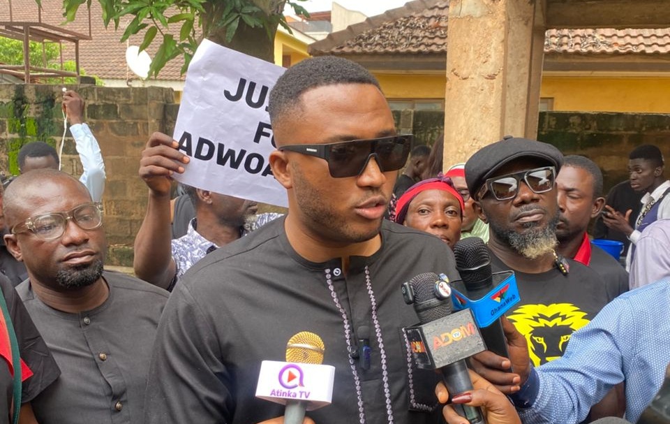 Man wearing dark outfit and sunglasses speaks to reporters at a protest, microphones in front of him.