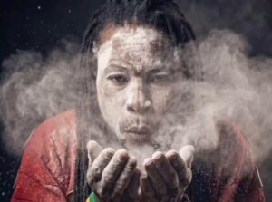 Man with a powdery face blowing dust from cupped hands toward the camera, wearing a red shirt.