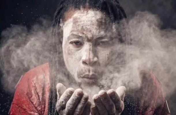 Man with a powdery face blowing dust from cupped hands toward the camera, wearing a red shirt.