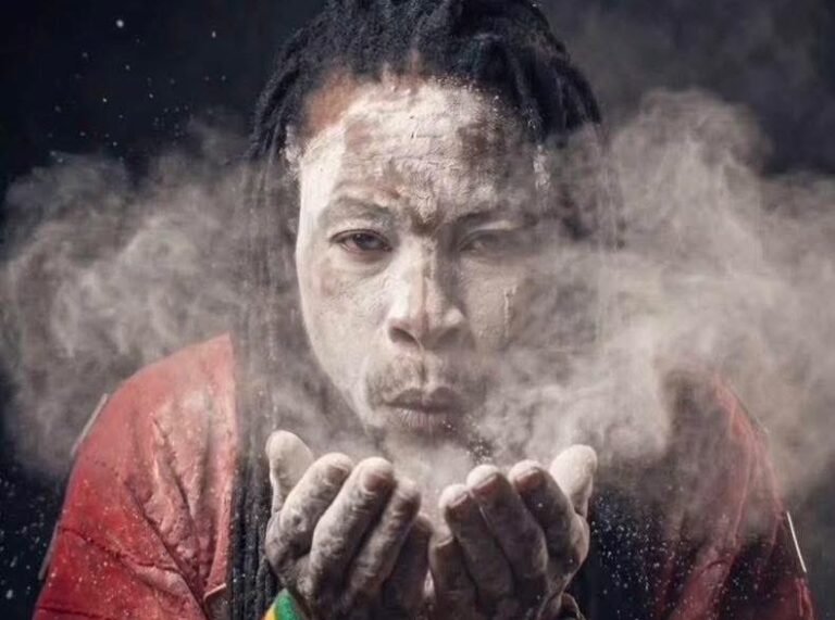 Man with a powdery face blowing dust from cupped hands toward the camera, wearing a red shirt.