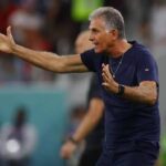 Athletic coach on a field gesturing with a left arm extended and right hand to chest, giving directions during a game.