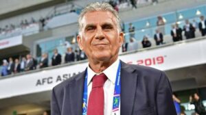 Smiling man in a navy suit and red tie with a lanyard at a sporting event, stadium stands in the background farsighted with banners.