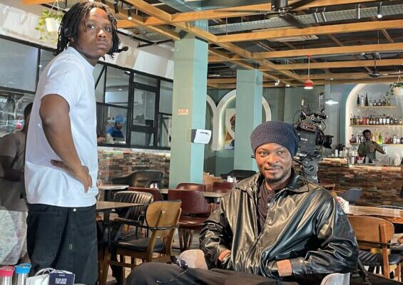 IMG_0319m-565x424 | Ghanaguardian.com Two men in a cafe: a standing man with dreadlocks in a light blue shirt beside a seated man in a black leather jacket and beanie, with a bar area in the background.