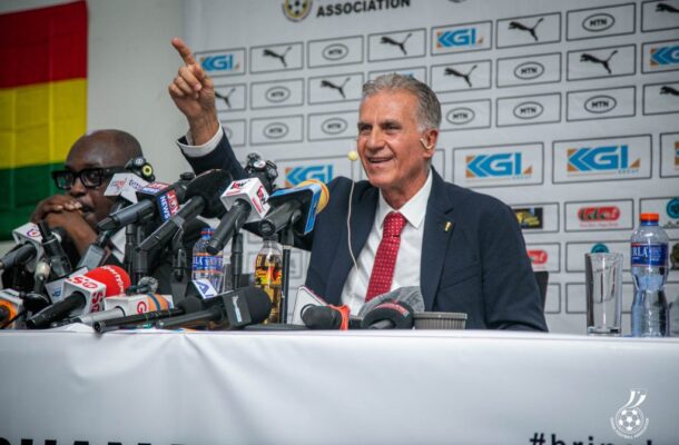 Man in a suit and red tie speaks at a press conference, raising his right hand beside numerous microphones.