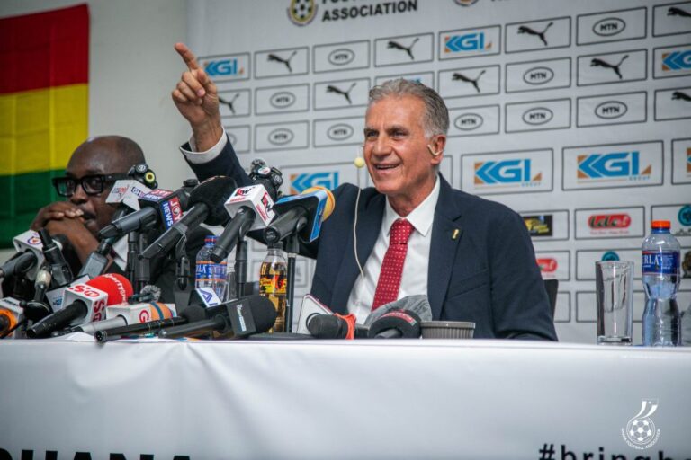 Man in a suit and red tie speaks at a press conference, raising his right hand beside numerous microphones.
