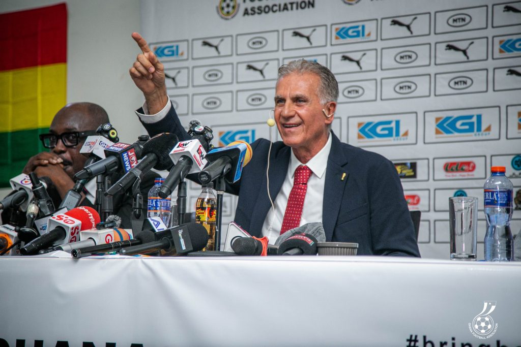 Man in a suit and red tie speaks at a press conference, raising his right hand beside numerous microphones.