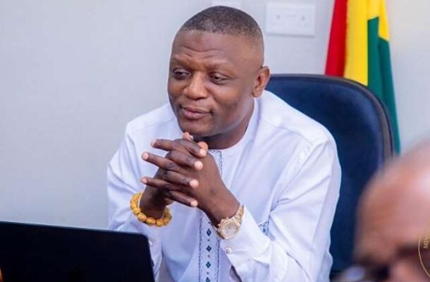 Man in a white traditional shirt sits at a desk with a laptop, hands clasped, in an office with a flag in the background.