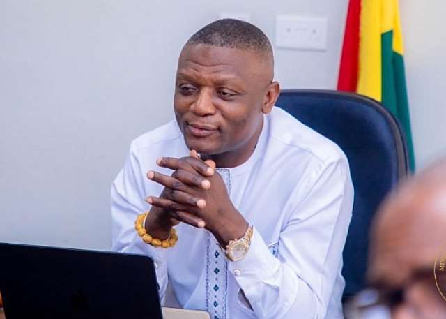 Man in a white traditional shirt sits at a desk with a laptop, hands clasped, in an office with a flag in the background.