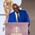 Man in a blue suit and glasses speaks at a podium during a Ghana World Cup fundraising launch event, with a large crest on the podium backdrop.