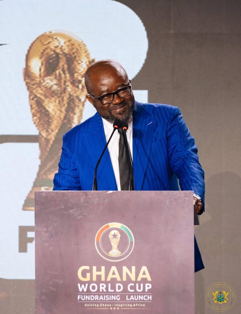 Man in a blue suit and glasses speaks at a podium during a Ghana World Cup fundraising launch event, with a large crest on the podium backdrop.