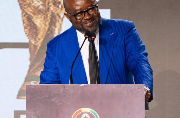 Man in a blue suit and glasses speaks at a podium during the Ghana World Cup Fundraising Launch event.
