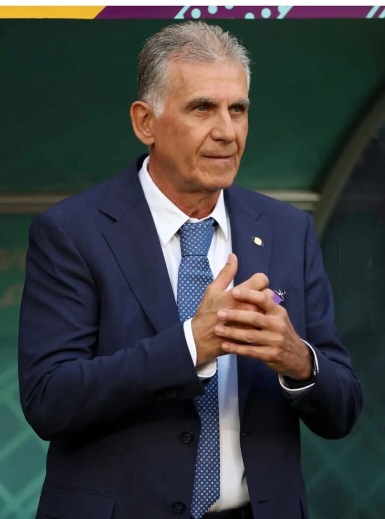 Man in a navy suit with white shirt and blue patterned tie, standing with hands clasped in front.