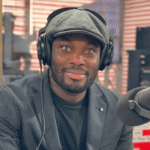 Man wearing a flat cap and large headphones, speaking into a microphone in a radio studio.