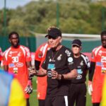 Coach in black giving instructions to a soccer team in red uniforms on a sunny training field, goal in background.