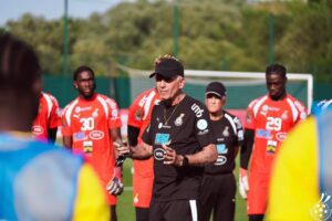 Coach in black giving instructions to a soccer team in red uniforms on a sunny training field, goal in background.
