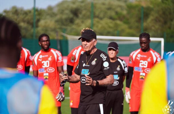 Coach in black giving instructions to a soccer team in red uniforms on a sunny training field, goal in background.