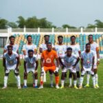 Team photo of eleven soccer players on a grassy field, goalkeeper in orange centered among teammates in light blue/white uniforms.