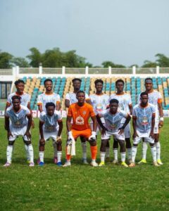 Team photo of eleven soccer players on a grassy field, goalkeeper in orange centered among teammates in light blue/white uniforms.
