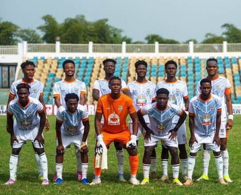 Team photo of eleven soccer players on a grassy field, goalkeeper in orange centered among teammates in light blue/white uniforms.