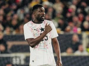 Soccer player in a white jersey with colorful geometric patterns touches his chest while standing on a crowded stadium field.