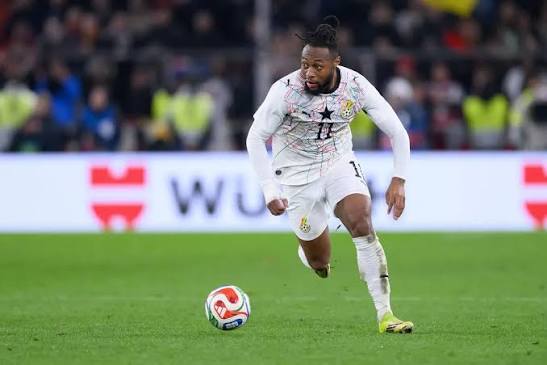 Soccer player in a white kit dribbles a ball on a green field during a match with a blurred crowd in the background.