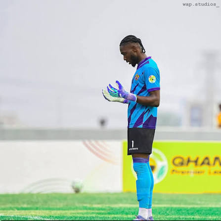 Soccer goalkeeper in a blue kit and purple gloves standing on the pitch, looking down at his hands with advertising boards in the background.