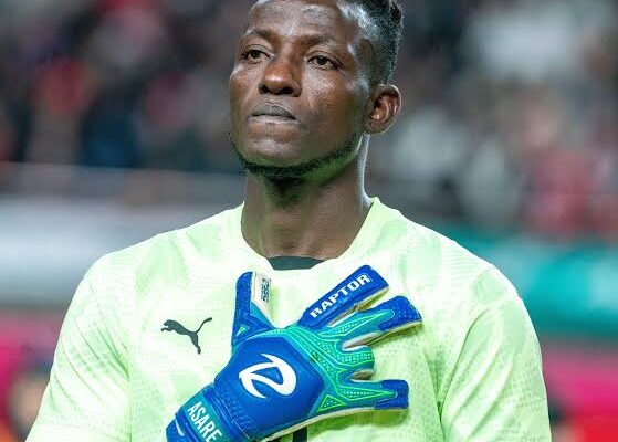 Soccer goalkeeper in neon green jersey with blue gloves, standing with hands crossed over chest in stadium.