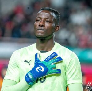 Soccer goalkeeper in neon green jersey poses with blue gloves, hand over heart, on the field during a match.