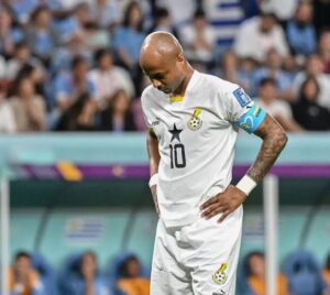 Soccer player in white jersey with number 10 stands with hands on hips on the field, stadium crowd in background, wearing captain's armband on left arm.