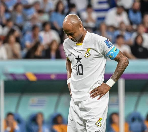 Soccer player in white jersey with number 10 stands with hands on hips on the field, stadium crowd in background, wearing captain's armband on left arm.