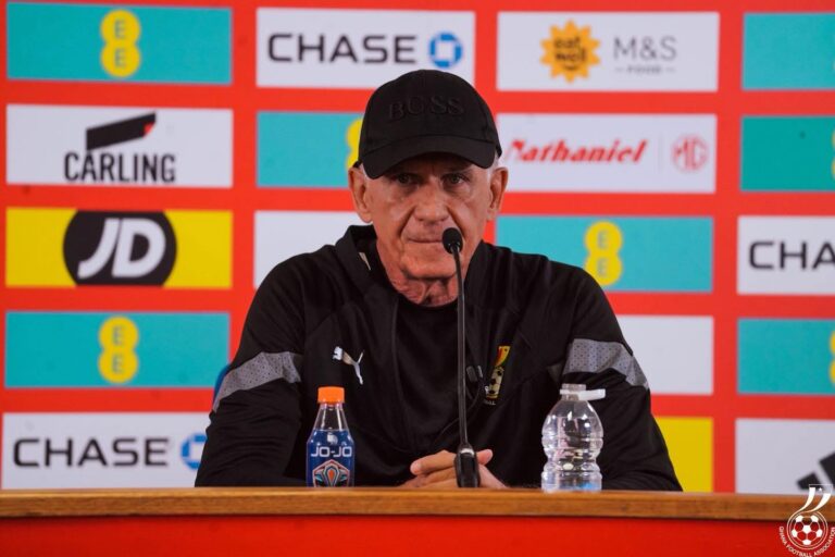 Man in a black cap and jacket speaks at a press conference, microphone in front, sponsor logos on a red backdrop behind him.