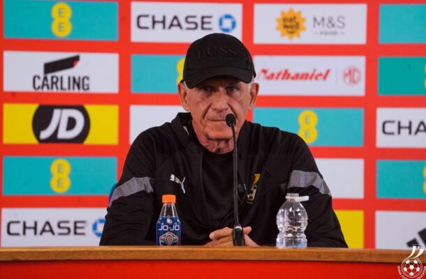 Older man in a black cap and jacket sits at a press conference table with a microphone, sponsor banners behind him.