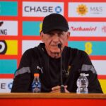 Man in a black cap and jacket sits at a press conference table with a microphone, sponsor banners behind him.