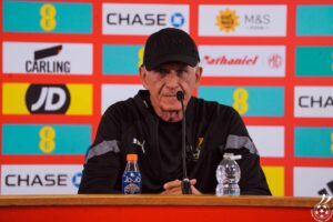 Male coach at a press conference wearing a black cap that says 'BOSS', speaking into a microphone with sponsor boards behind him.