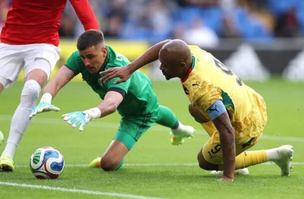 Soccer scene with a green-clad goalkeeper diving to reach the ball while a yellow-clad player slides in, and a red-clad opponent's leg is visible nearby.